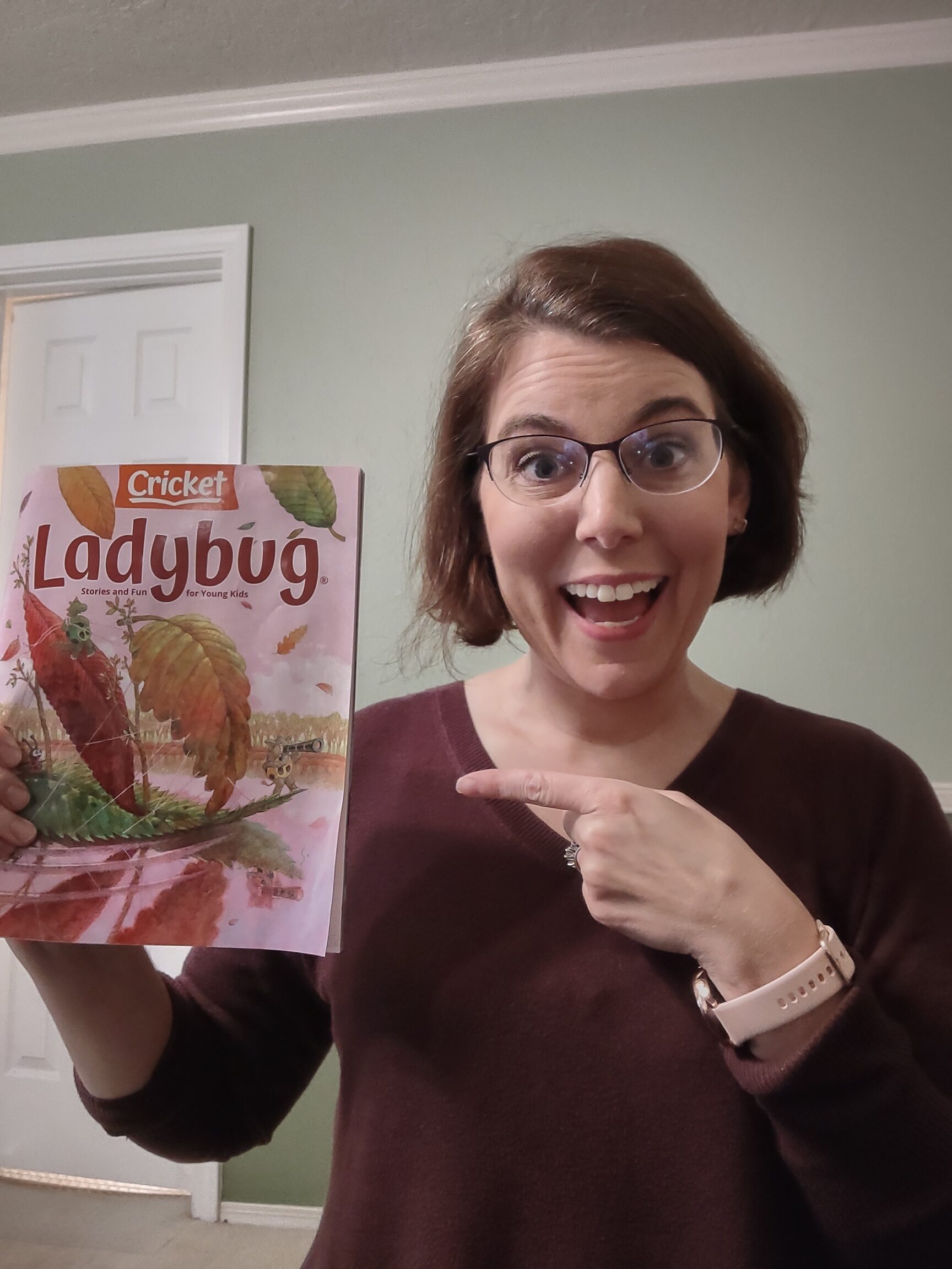 Susan, a white woman with short brown hair and glasses, holds up a copy of Ladybug Magazine and points at it with a wide smile.