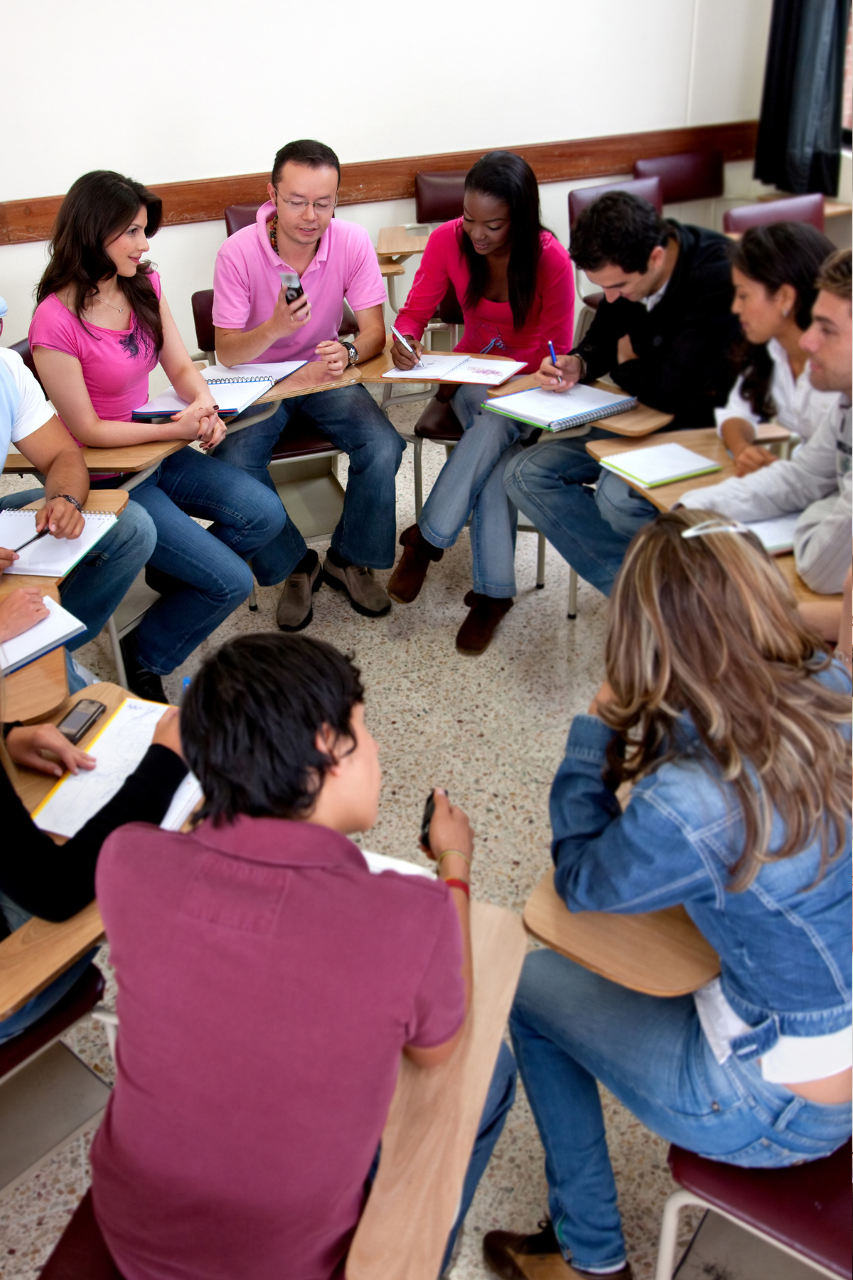 A group of high school or college age students sit at desks arranged in a circle with writing implements and papers on them.