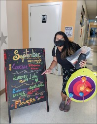 A woman with black hair, brown skin, and black clothing holding an armful of activities, including a velcro bean bag toss, points to a list of activities on a blackboard sign, one of which reads "Library Lady"