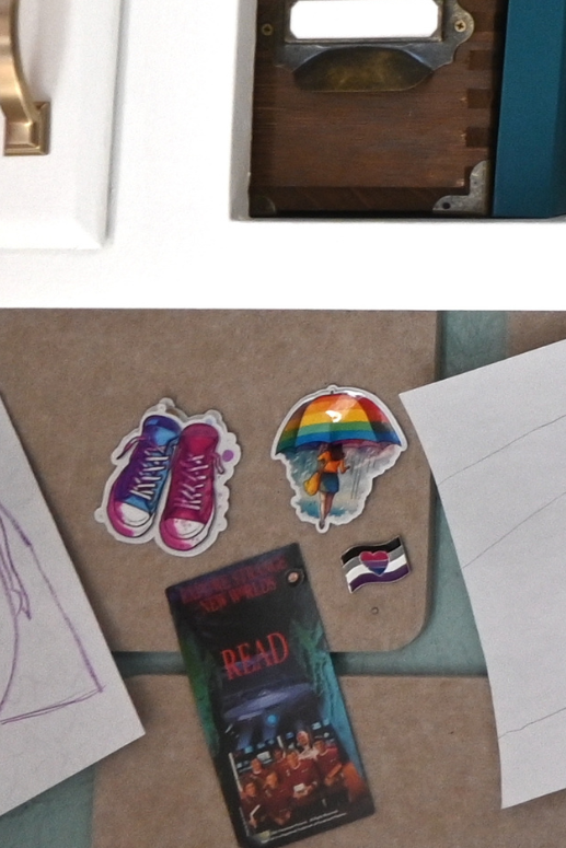 A desk with white cabinets with brass handles and a bulletin board with a crayon drawing of a girl's face smiling, a vinyl sticker of chuck taylors in bi-pride colors, a vinyl sticker of a girl standing under a rainbow umbrella, a biromantic ace pride pin, papers, a teal notebook, and a plant.