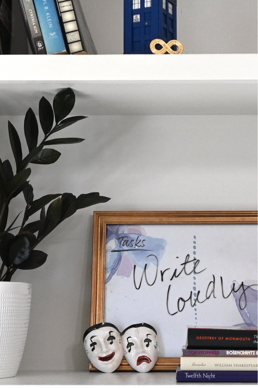 White bookshelves with writing and library science books, a houseplant, ceramic theatre masks, a stack of books, a wooden TARDIS, a neurodivergent pride pin, and a picture frame/white board reading "write loudly"