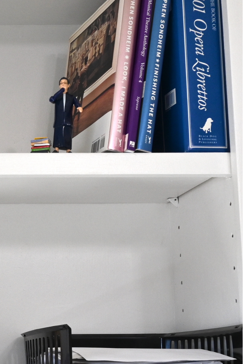 Books about musical theatre and opera next to a librarian action figure on one bookshelf and below a stack of papers, a stack of books, and a Star Trek TNG communicator.