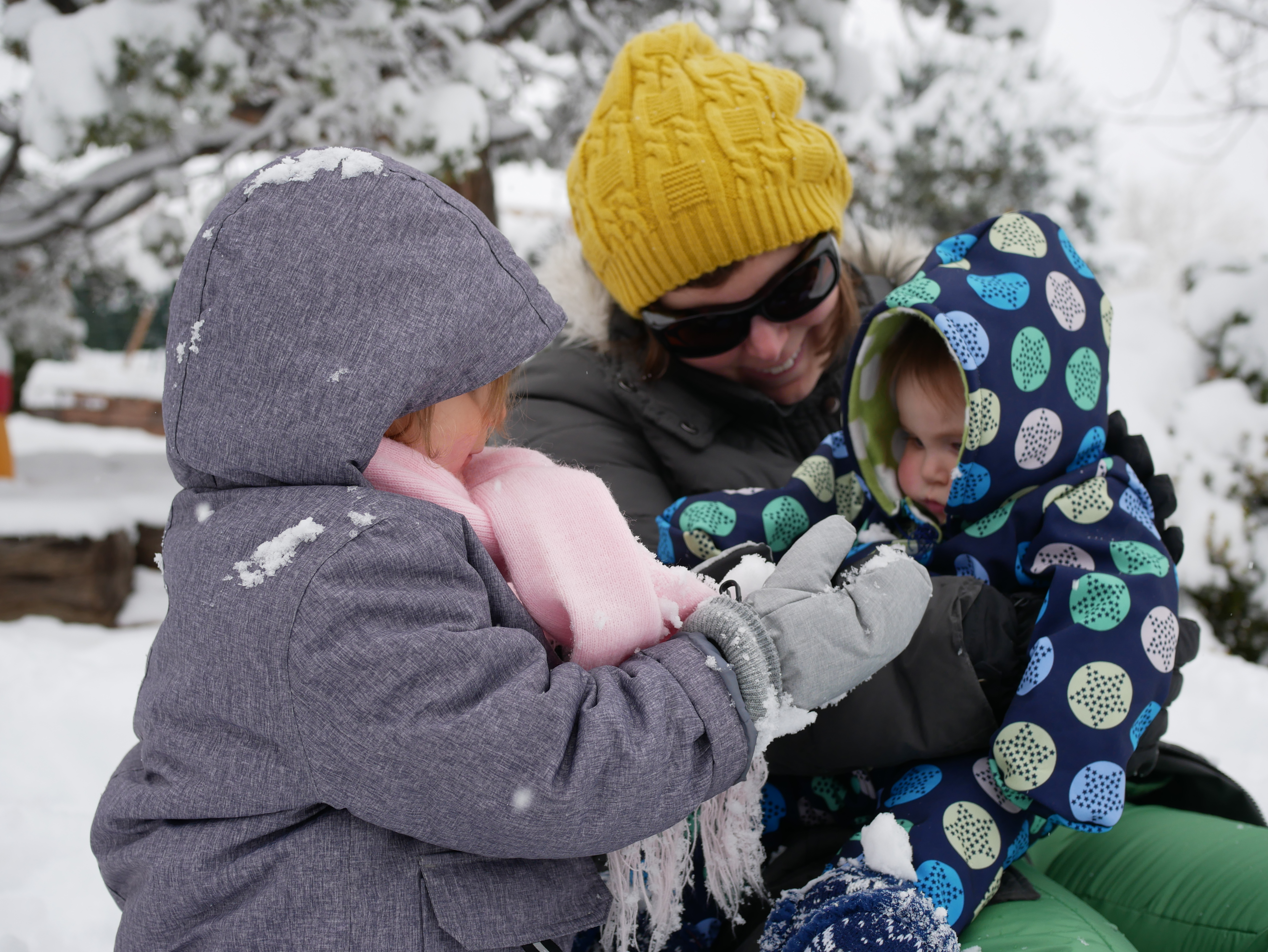A smiling mom in a yellow winter hat and sunglasses holds an infant in a snow suit and while another child bundled up in a gray winter coat and pink scarf offers the baby a handful of snow.